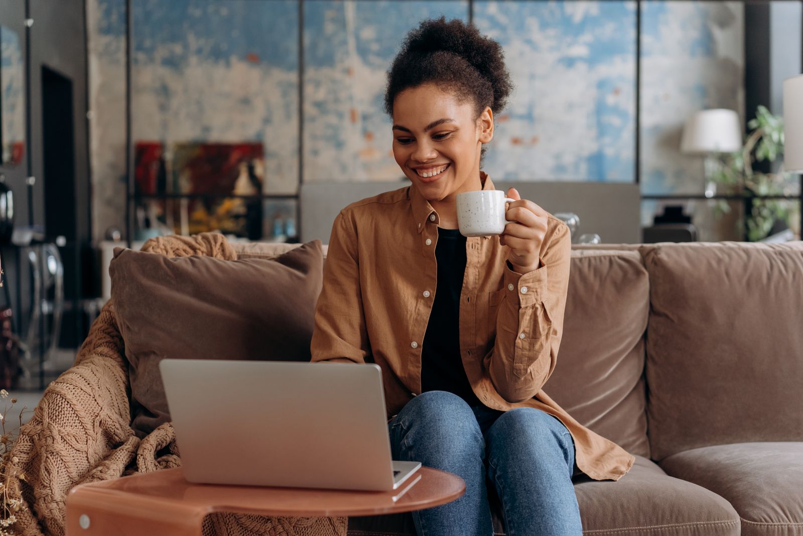 A smiling woman relaxing on a sofa with coffee and an open laptop